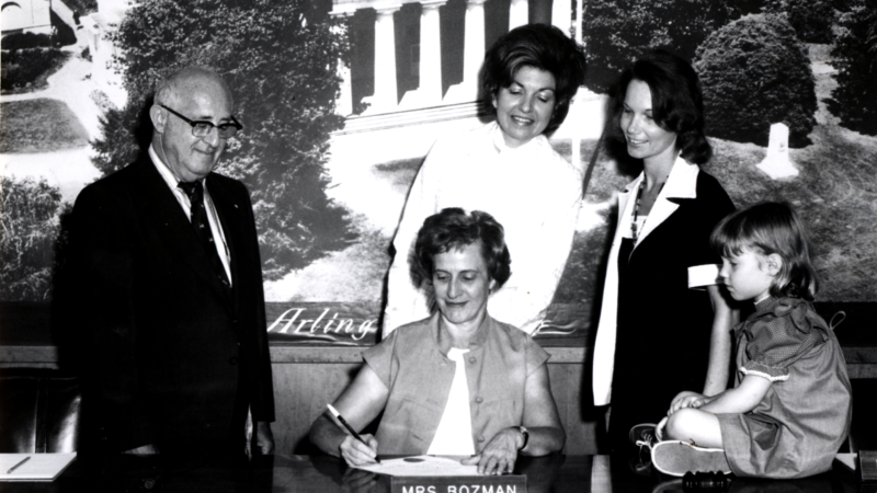 Ellen Bozman working in the County Board office, surrounded by an unidentified man, two women and a child sitting on the desk.
