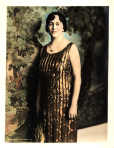 Full length portrait of Ruby Lee Minar in gold sequined dress standing in front of a background of green foliage. Photo taken by Underwood & Underwood, Inc. Washington, D.C., 1930s