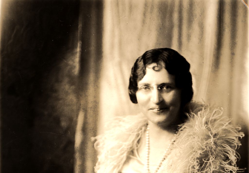 Seated portrait of Ruby Lee Minar wearing a cape with a feather collar and long pearl necklace. Photo taken by Harris & Ewing, Washington, D.C.